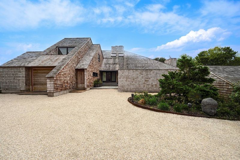 The approach: the low, cedar-shingled Jaffe house with its long sloping roofs and stone-clad chimney mass, seen across a wide gravel motor court. at 95 Jobs Lane, Bridgehampton