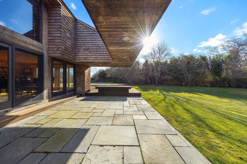 A bluestone terrace under a dramatic cantilevered roof overhang, the sun low over the lawn and tree line. at 95 Jobs Lane, Bridgehampton — photo 7 of 12
