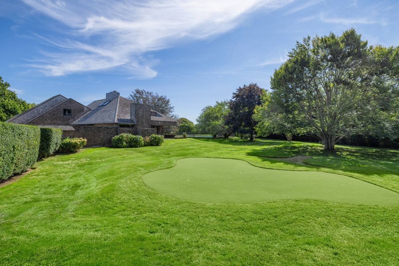 The putting green on the lawn with the house beyond, framed by hedges and a specimen tree. at 95 Jobs Lane, Bridgehampton — photo 11 of 12